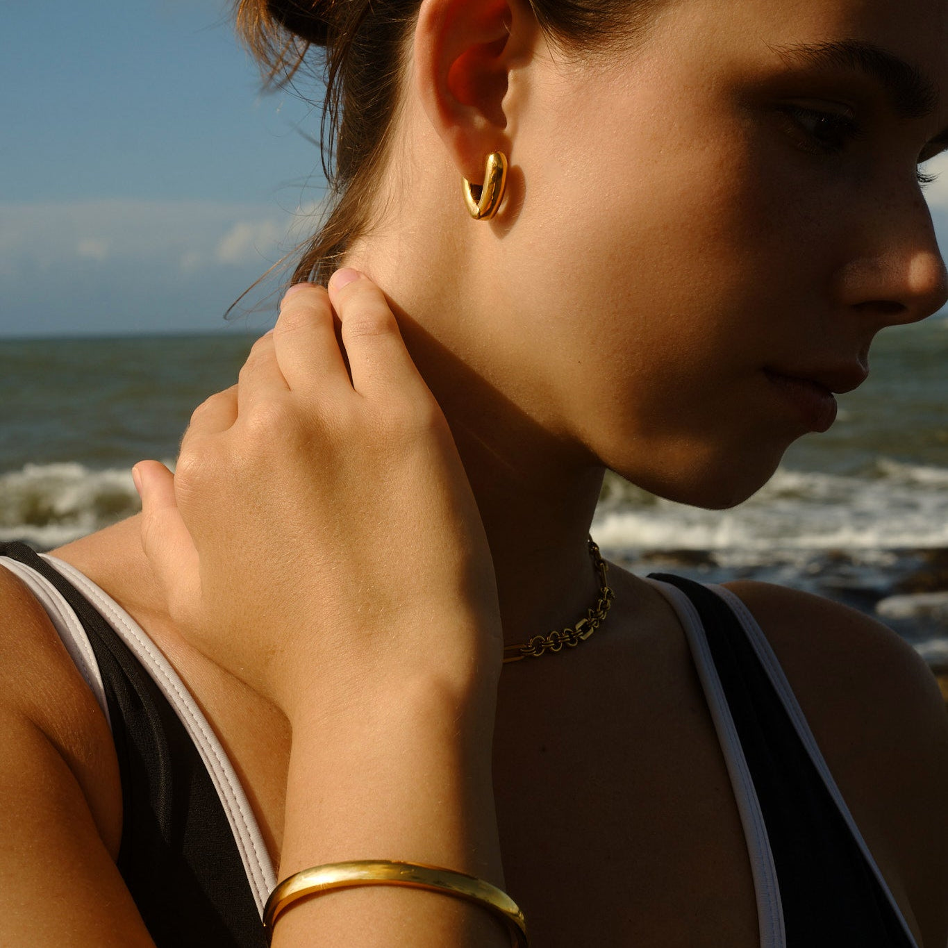Woman adjusting her gold earring on a beach with ocean and sky in the background