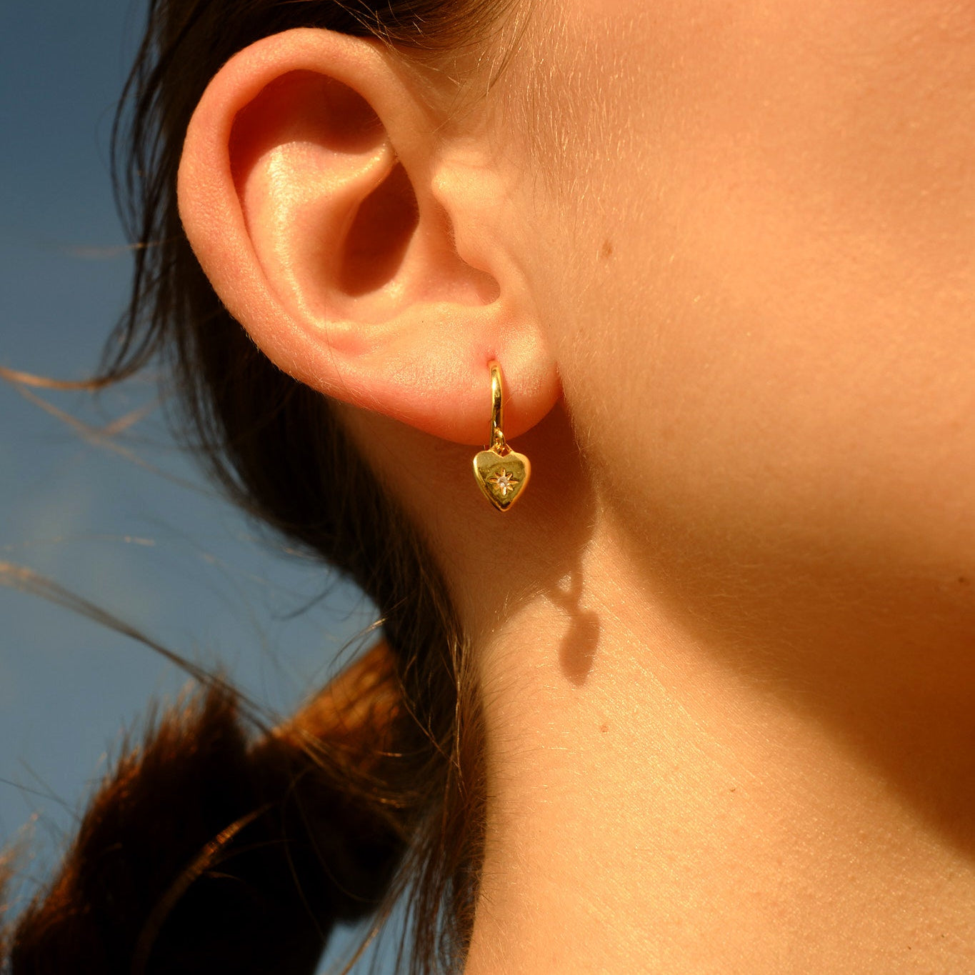 Close-up of a person wearing a gold heart-shaped earring with a clear blue sky background.