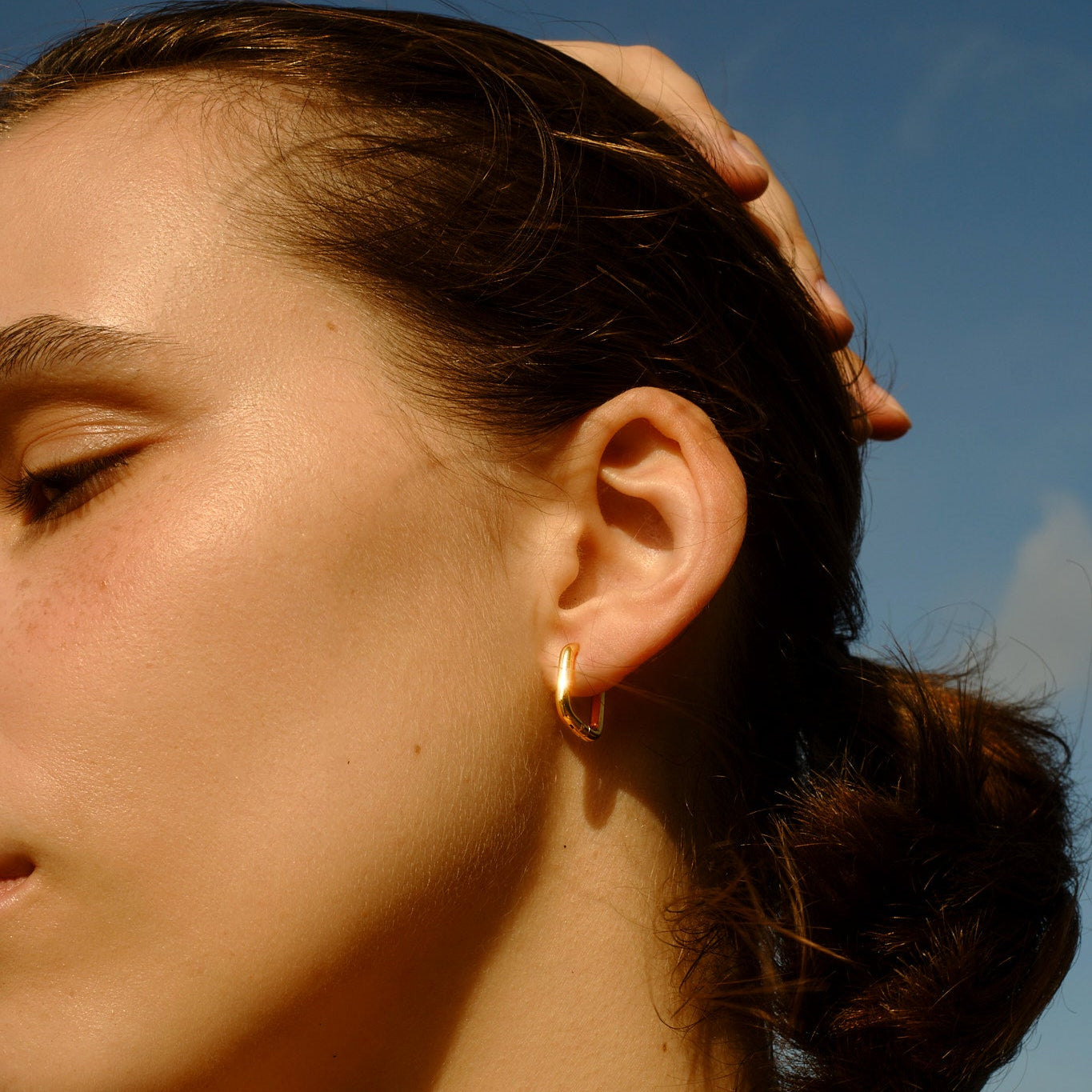 Close-up of a woman's ear with a square earring against a blue sky.