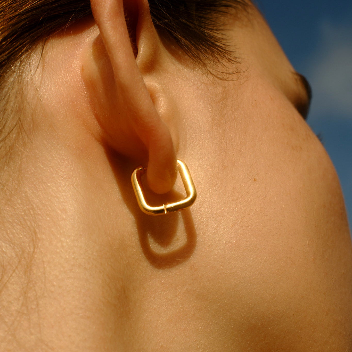 Close-up of a square gold hoop earring worn by a person with a blurred background